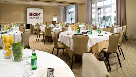Large meeting room room with nine tables neatly decorated with white tableclothes and topped with waters, fresh fruit, and Hershey bars on the tables.