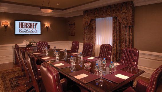 Dimmed meeting room with one large round table with comfortable office chairs lined, the table topped with waters, candies, and notepads at each seat.