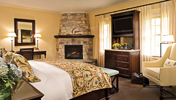 King size bed in the center of the room surrounded by lit fireplace, beside table with two lit lamps, a vanity, and a chair in the corner.