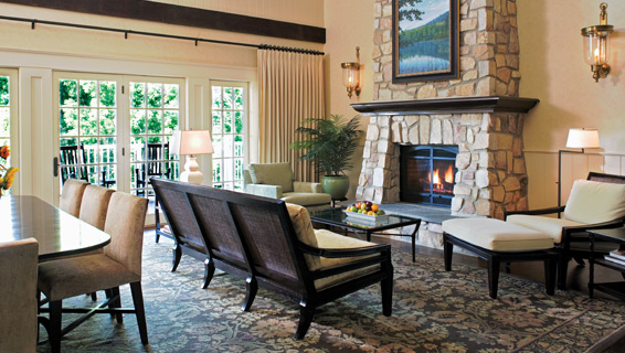 Room with filled natural sunshine shining through three glass doors, a giant fireplace placed in the middle and surrounded by comfortable seating and dining table.