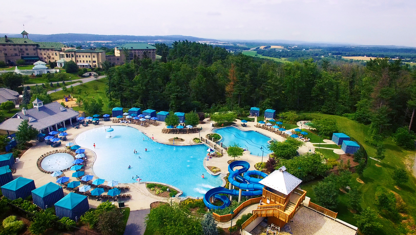drone view of hotel guests enjoying the tree-lined hotel outdoor pool that overlooks the town of Hershey, PA. Guests seen enjoying the waterslide, rows of seating incuding private cabanas and lounge chairs just inches away from the pools ledge, and giant hot tub that is located off of pool main entrance.