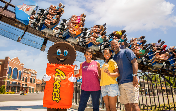 Family of three posing with hersheypark character, Reese's, in front of Candymonium coaster with riders passing by in the background. 