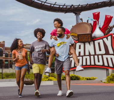 Dad giving his wife a piggy-back ride while walking alongside his two children, who are holding up hersheypark milkshakes, with Candymonium coaster and Candymonium sign behind them.