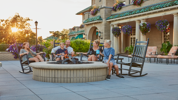 Family of five sitting in rocking chairs surrounding the hotel firepit, making s'mores and engaging in joyful conversation.