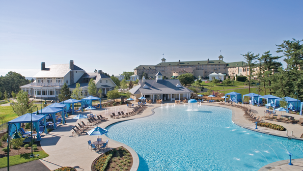 The clear blue hershey hotel outdoor pool, secluded behind the hotel, is surrounded by various seating, including cabanas, picnic tables with umbrellas, and lounge chairs close to the water's edge.