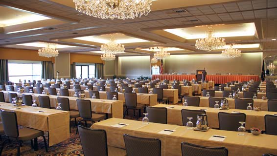 Extremely large meeting room lined with over 15 long tables, lined with chairs, facing the front of the room where a stage and potium is set up for presentation.