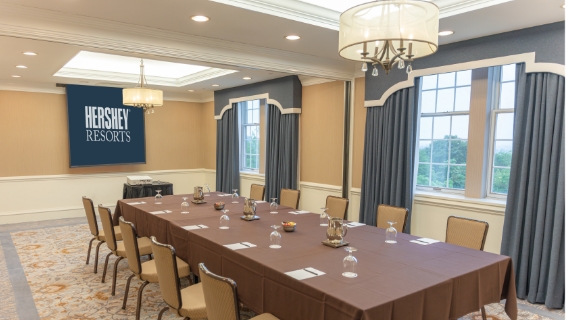 Extremely large meeting room lined with over 15 long tables, lined with chairs, facing the front of the room where a stage and potium is set up for presentation.