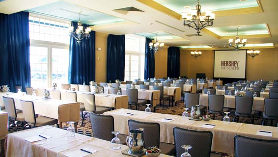 Medium-sized meeting room with blue curtains hung by large windows, rows of long tables, and a presentation screen situated at the front of the room.